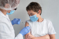 Coronavirus, flu or measles vaccine concept. Medic, doctor, nurse, health practitioner vaccinates teenage boy with vaccine in syringe. She is wearing uniform, hut and gloves. Both have face mask